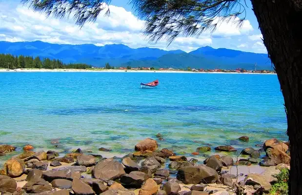 Mar calmo e pedras da Praia do Sonho com montanhas ao fundo, em Palhoça